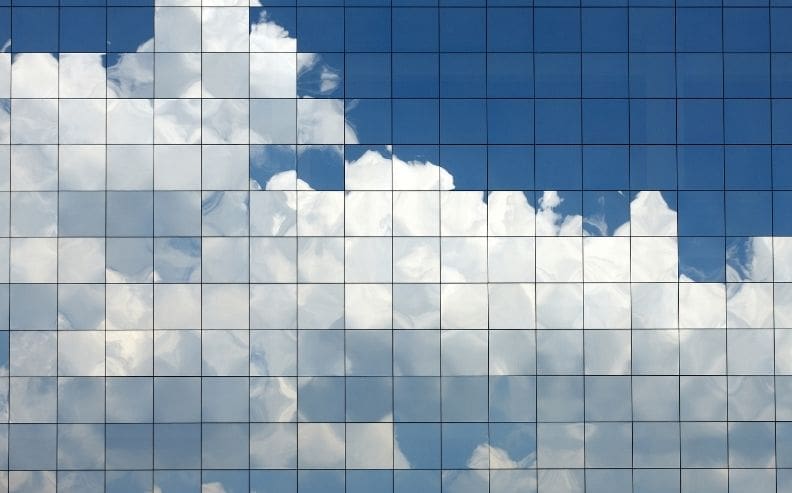 Reflection of blue sky and white clouds on a glass building’s grid-like window panels, creating a geometric pattern with sections of sky and clouds.