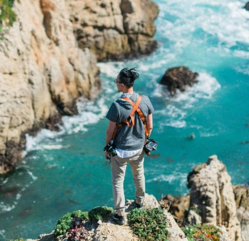 A person wearing a camera harness stands on a rocky cliff edge overlooking turquoise ocean water below, with rugged rocks and waves in the background.