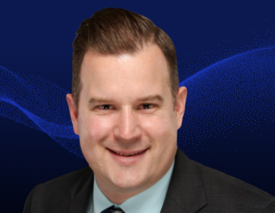 A man with short brown hair, wearing a dark suit, light blue shirt, and a tie, smiles at the camera against a dark blue background with abstract wave patterns.