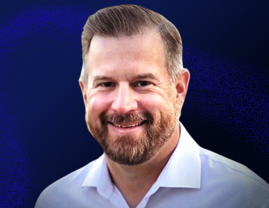 A smiling man with short brown hair and a beard, wearing a white collared shirt, is shown against a dark blue background.