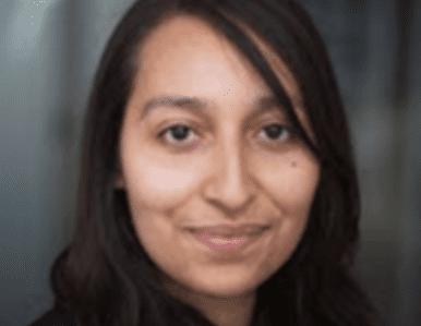 A person with long, dark hair and brown eyes smiles softly at the camera. The background is blurred, keeping the focus on their face.