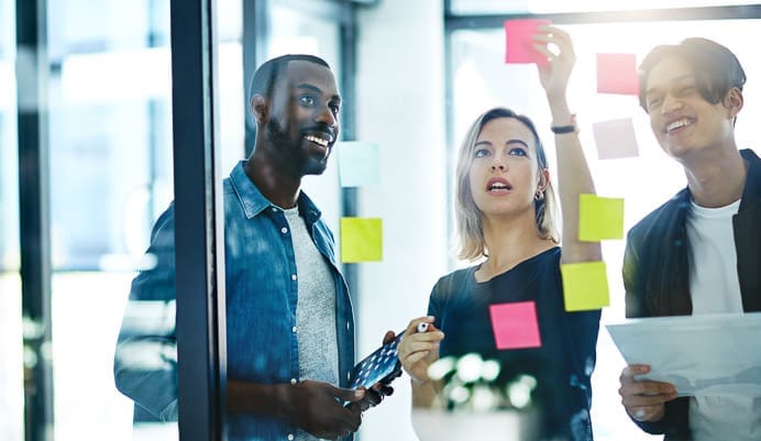 Three people stand together in an office, brainstorming and placing colorful sticky notes on a glass wall. One person holds a tablet, another writes on a note, and all appear engaged and focused on the discussion.