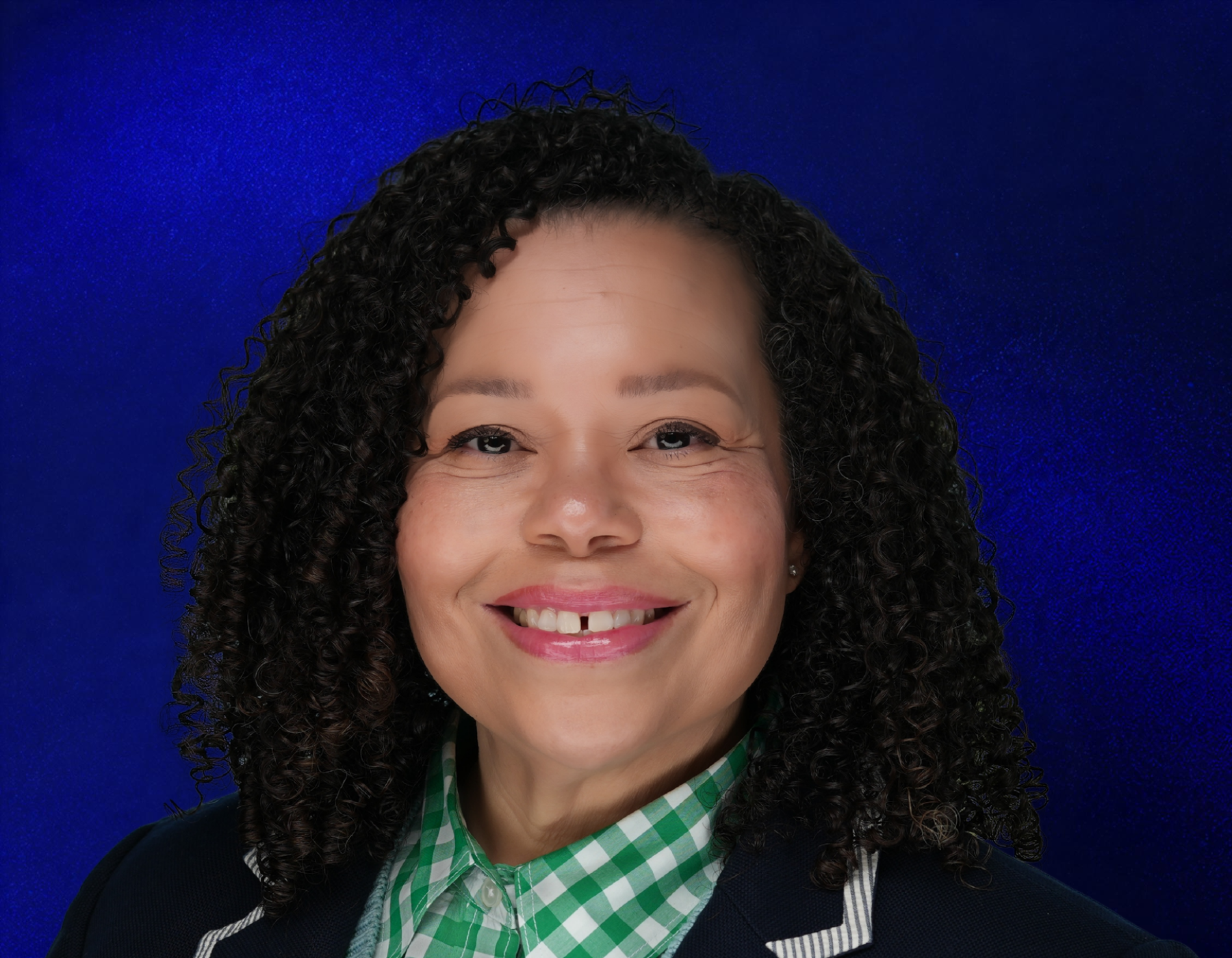 Smiling woman with curly hair wearing a green checkered shirt and a dark blazer with white trim, posed against a vibrant blue background.