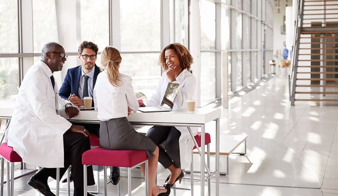 Four professionals, two in white lab coats and two in business attire, sit around a table in a modern, sunlit office with large windows, engaged in a discussion.