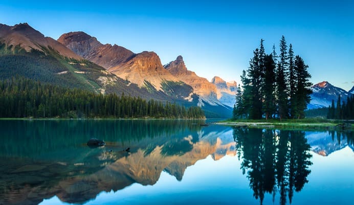 A serene lake reflects tall evergreen trees and rugged, sunlit mountains under a clear blue sky, mirroring the harmony and security needed for robust Identity for Critical Infrastructure.