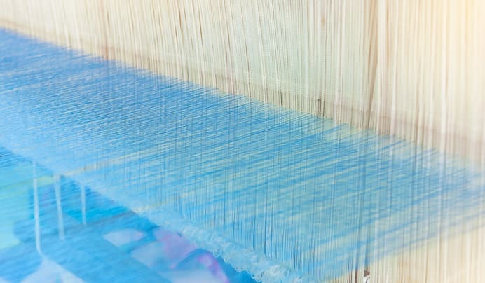 Close-up of light blue and beige threads stretched vertically and horizontally on a loom, illustrating the weaving process in textile production. The threads form an intricate, organized pattern.