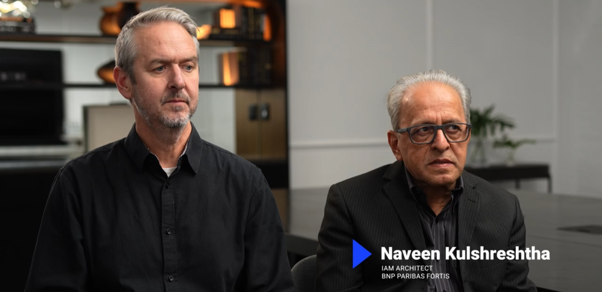 Two men sit side by side in a modern office. The man on the right, identified as Naveen Kulshreshtha, IAM Architect at BNP Paribas Fortis, wears glasses and a dark suit. The other man has gray hair and wears a black shirt.