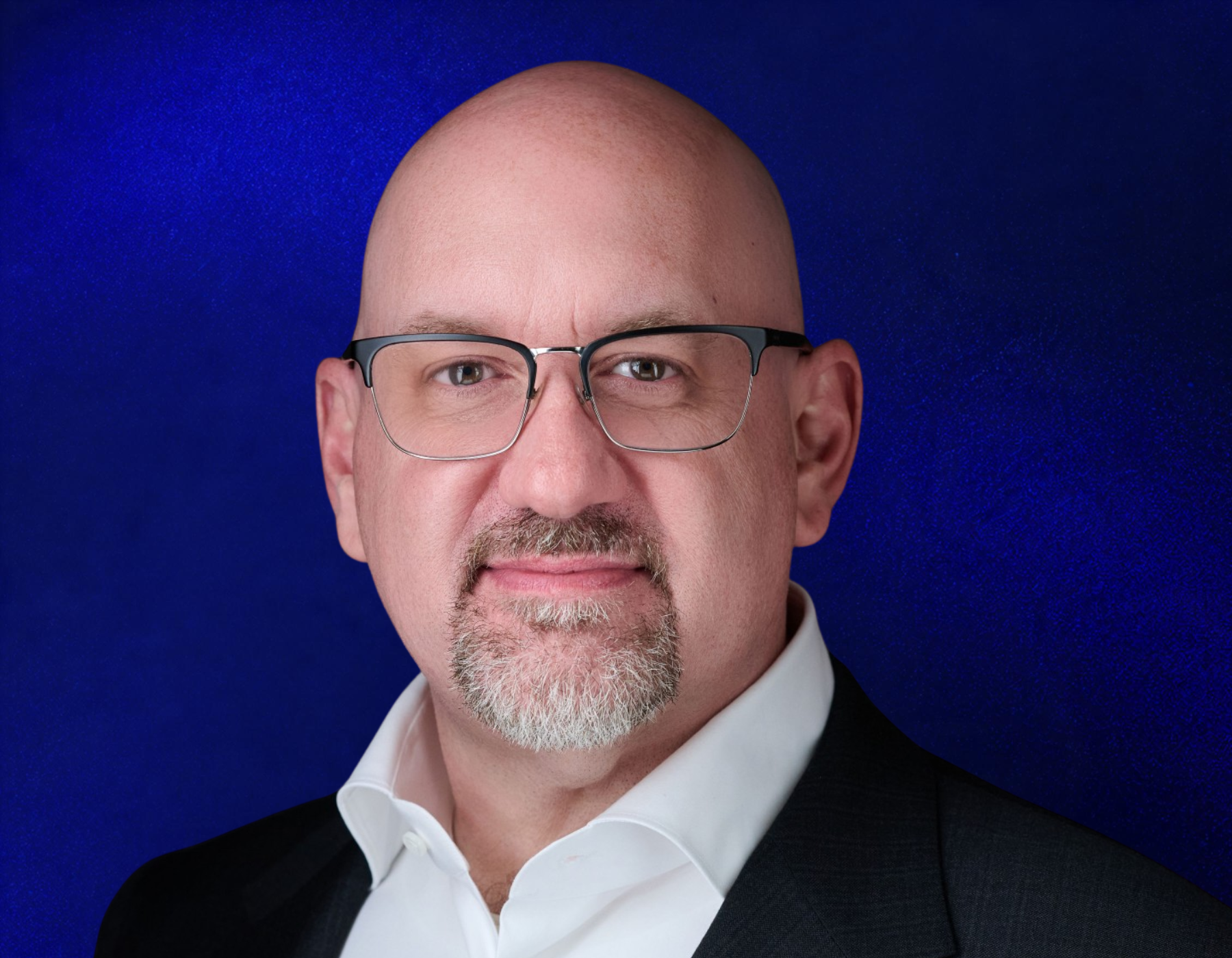 A bald man with glasses, a goatee, and mustache, wearing a dark suit and white shirt, poses against a solid blue background.