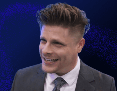 A man with styled brown hair, wearing a grey suit, white shirt, and patterned tie, smiles while looking slightly to the side against a dark blue background.