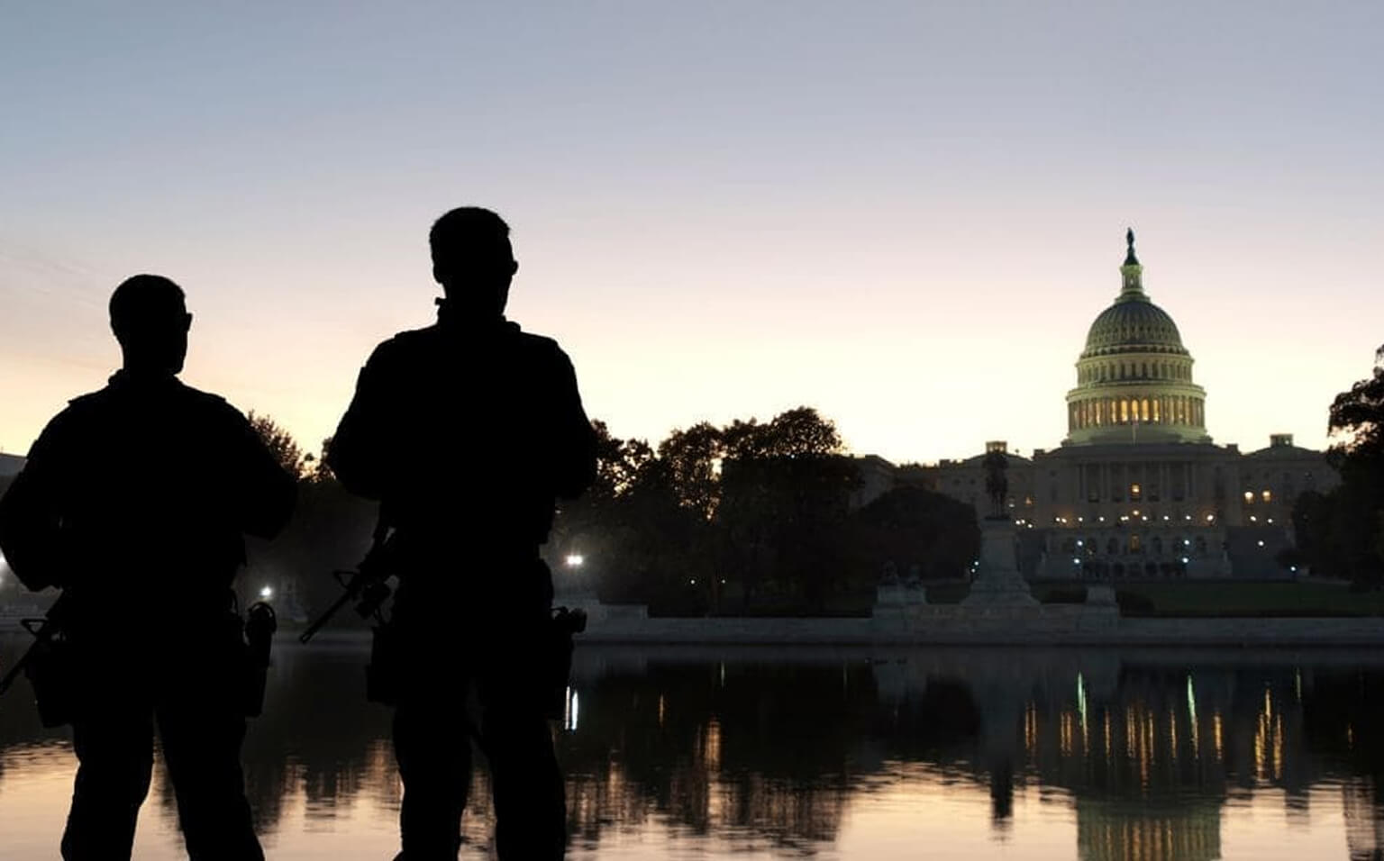 Two silhouetted security officers stand with rifles near a reflective pool at dusk, facing the illuminated United States Capitol building against a dimly lit sky.