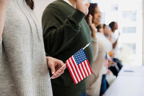 A group of people stand in a row with raised right hands, taking an oath. One person in the foreground holds a small American flag. The scene suggests a citizenship or naturalization ceremony.