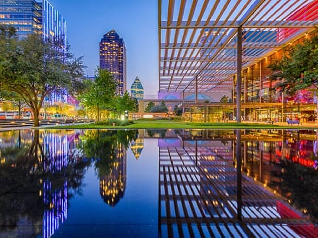 Colorful, modern cityscape at dusk with glass buildings and trees reflecting in a large, still water feature; vibrant lights and a partially open metal canopy create an urban, lively atmosphere.