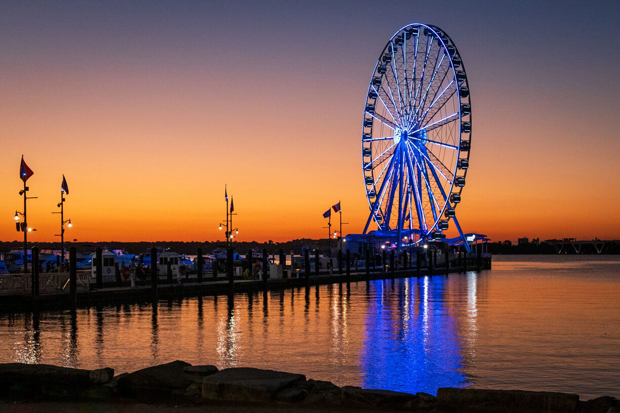 A large, illuminated Ferris wheel stands on a pier at sunset, reflecting blue lights on the calm water. Flags line the pier, and the sky glows orange and purple above the distant shoreline.