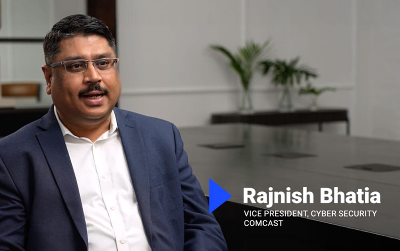 A man in a suit and glasses sits in an office, discussing identity data. On-screen text reads: Rajnish Bhatia, Vice President, Cyber Security, Comcast. A plant and conference table are in the background.