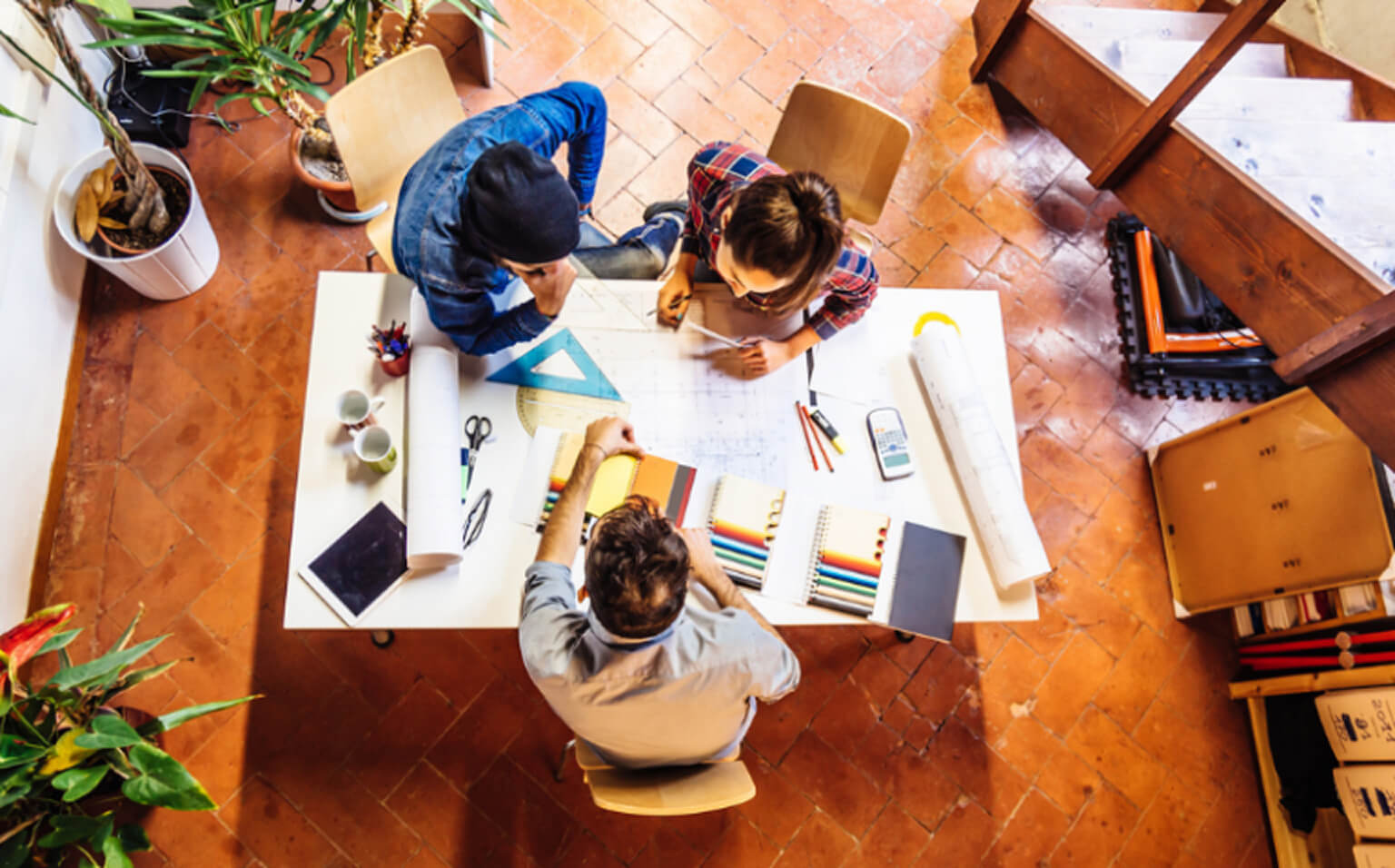 Three people sit around a white table with papers, stationery, and a laptop, collaborating on a project in a bright room with a tiled floor and plants, viewed from above.