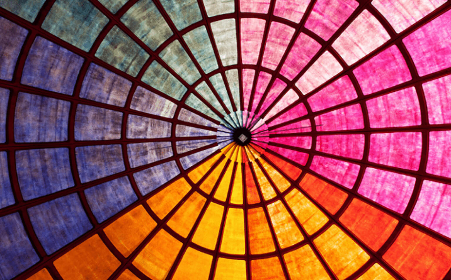Colorful stained glass ceiling seen from below, forming a circular pattern with radiating segments in shades of blue, red, yellow, pink, and purple, converging at a central point.