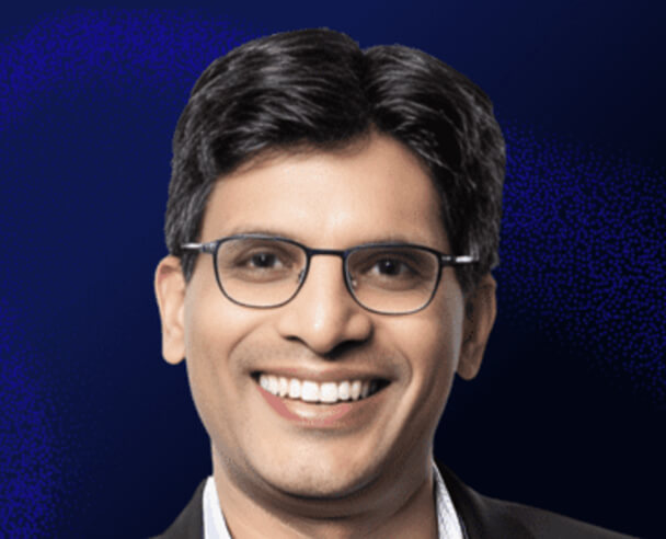 A man with short, dark hair and glasses is smiling, wearing a dark suit jacket over a light shirt, with a dark blue gradient background.