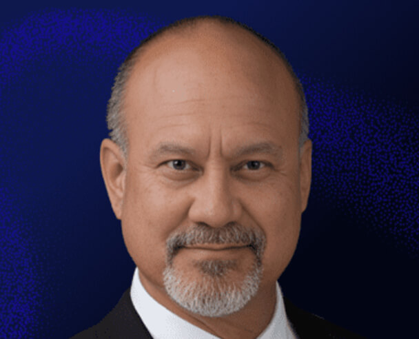 A middle-aged man with a bald head, gray goatee, and mustache is wearing a dark suit and white shirt, posing against a dark blue background.