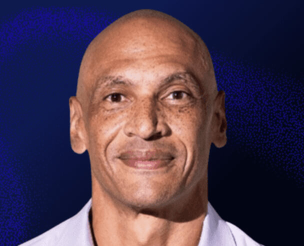 A middle-aged man with a shaved head, light brown skin, and freckles smiles gently at the camera against a dark blue background.
