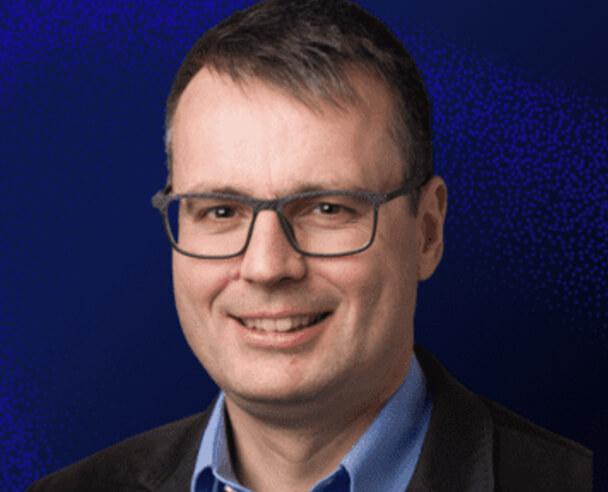 A man with short dark hair and glasses, wearing a blue collared shirt and dark blazer, smiling in front of a blue textured background.