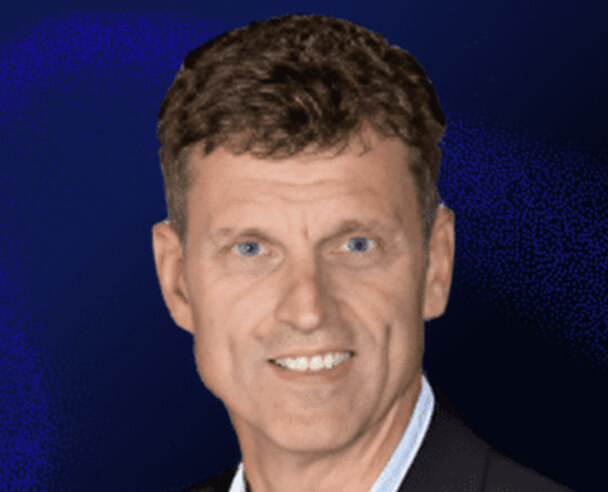 A middle-aged man with short brown hair and blue eyes is smiling, wearing a dark jacket and light blue collared shirt, in front of a dark blue background.