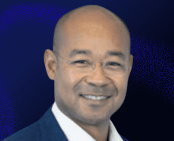 A smiling man with a bald head, wearing glasses and a dark suit jacket over a white shirt, is pictured against a dark blue background.