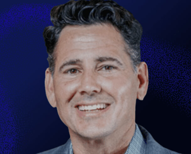 A man with dark, wavy hair and light skin is smiling and wearing a checkered shirt and jacket against a dark blue background.