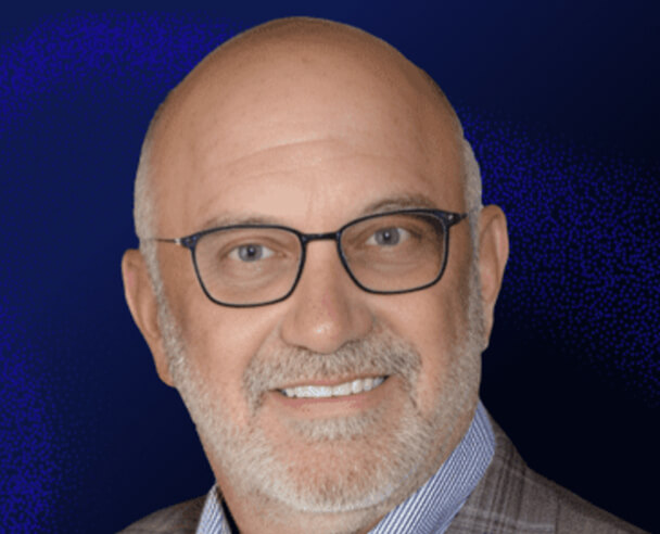 A middle-aged man with a bald head, gray beard, and glasses smiles at the camera. He is wearing a plaid jacket and a collared shirt, set against a dark blue background.