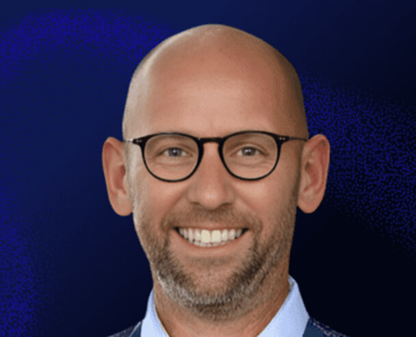 A smiling bald man with glasses and a short beard, wearing a blue shirt and jacket, is pictured in front of a dark blue background.