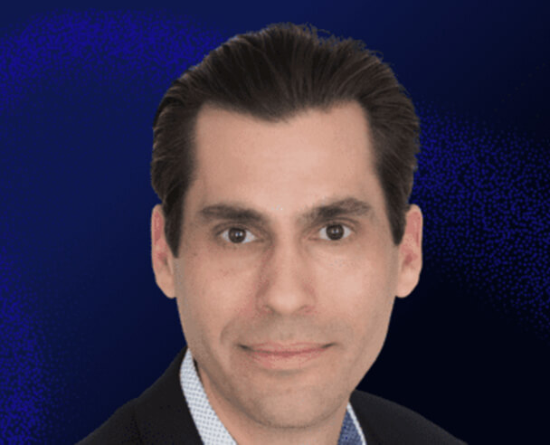 A man with dark, slicked-back hair wearing a dark suit jacket and a light-colored shirt, set against a dark blue gradient background.