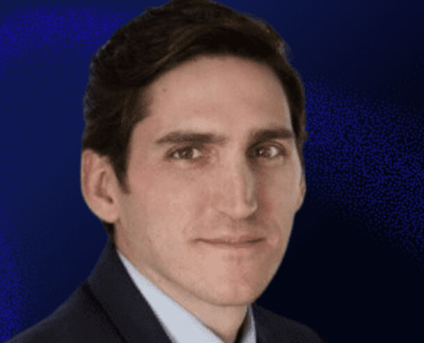 A man with short dark hair wearing a suit jacket and light blue shirt, looking at the camera with a neutral expression. The background is a dark blue gradient.