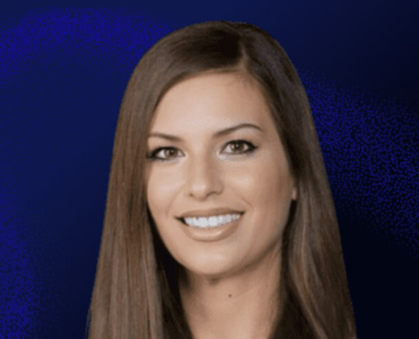A woman with long straight brown hair and light skin smiles at the camera against a dark blue background.