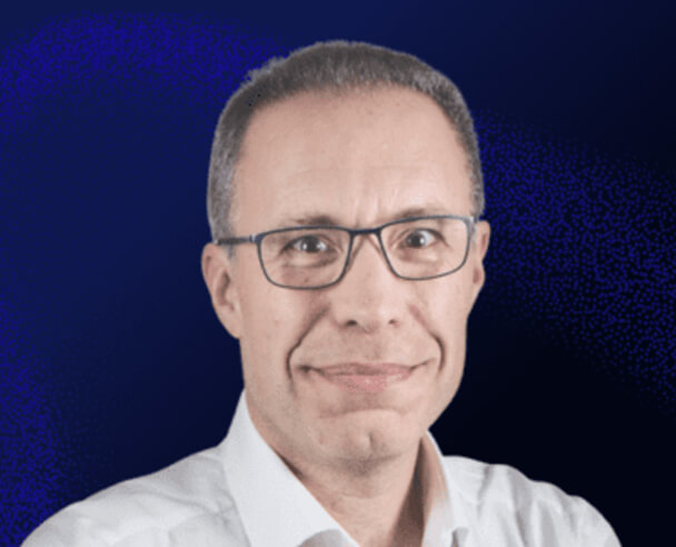 A middle-aged man with short gray hair and glasses smiles at the camera. He is wearing a white shirt and is posed against a dark blue background.