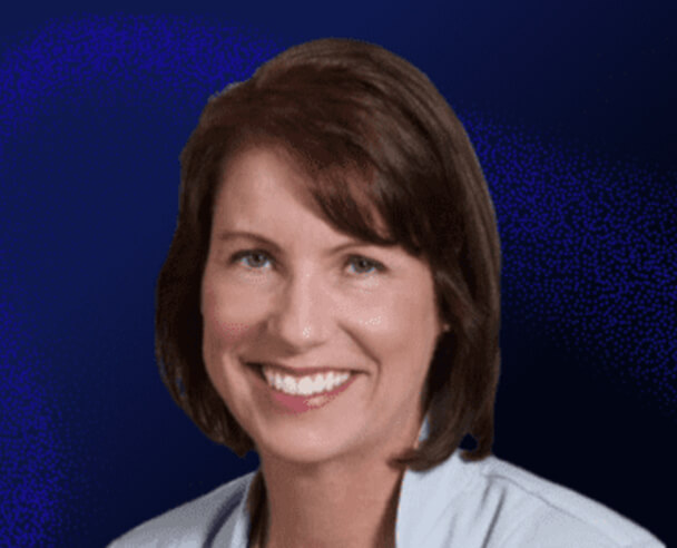 A woman with short brown hair and a light blue collared shirt smiles in front of a dark blue background.