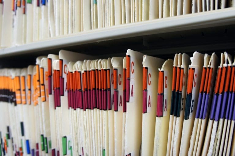 Rows of paper file folders on a shelf, each with colored index tabs and letters, organized vertically and tightly packed together, representing an organized filing or record-keeping system.