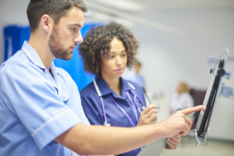 Two healthcare professionals, one male and one female, wearing scrubs and stethoscopes, look at a screen. The man points at the screen while the woman holds a clipboard. The setting appears to be a hospital.