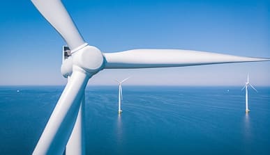 Close-up of a large white wind turbine at sea, with two more wind turbines visible in the distance against a clear blue sky and calm ocean water.