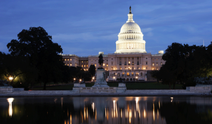 The U.S. Capitol building illuminated at dusk, with its reflection visible in the water in the foreground and trees framing the scene.