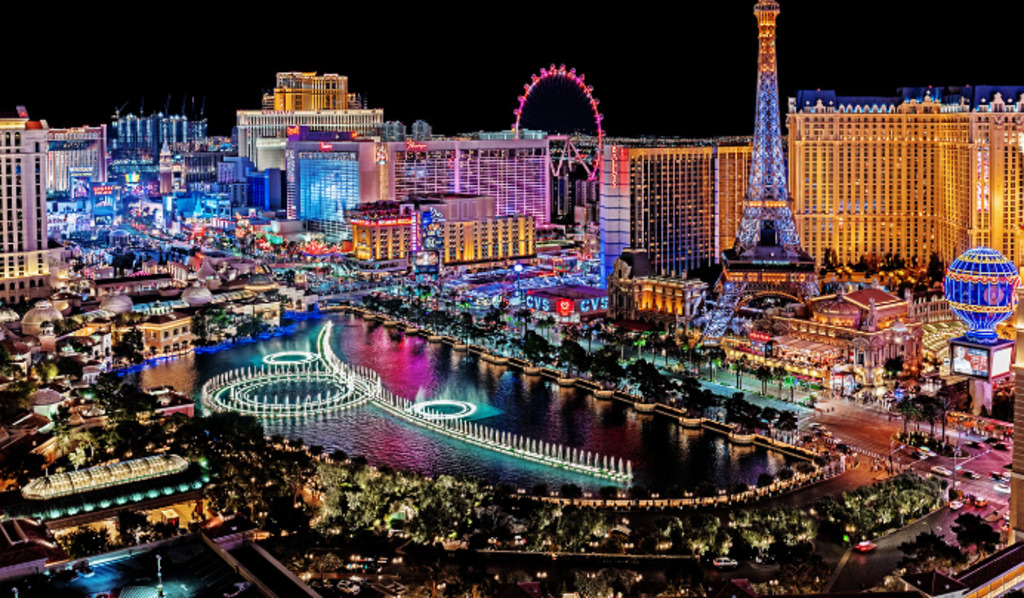 A vibrant nighttime view of the Las Vegas Strip, featuring illuminated hotels, casinos, a large fountain display, the Eiffel Tower replica, and the High Roller Ferris wheel against a dark sky.