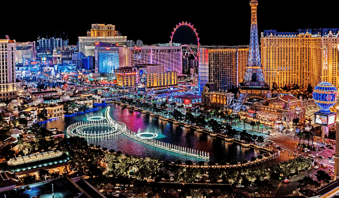 A vibrant nighttime view of the Las Vegas Strip featuring brightly lit hotels, casinos, the Eiffel Tower replica, a Ferris wheel, and colorful fountains in front of the Bellagio hotel.