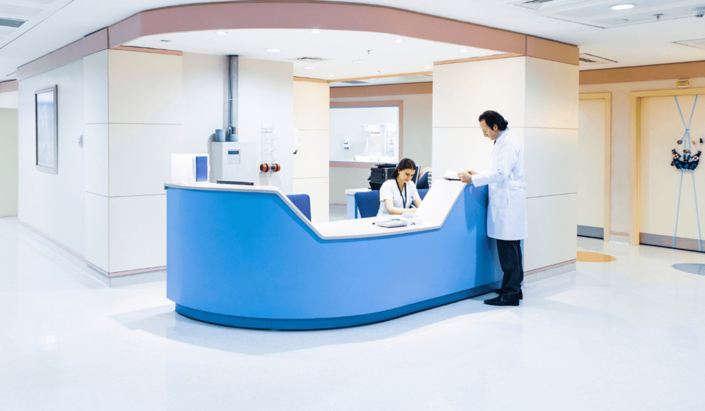 A doctor in a white coat stands at a blue and white hospital reception desk, speaking with a seated receptionist in a clean, modern medical facility.