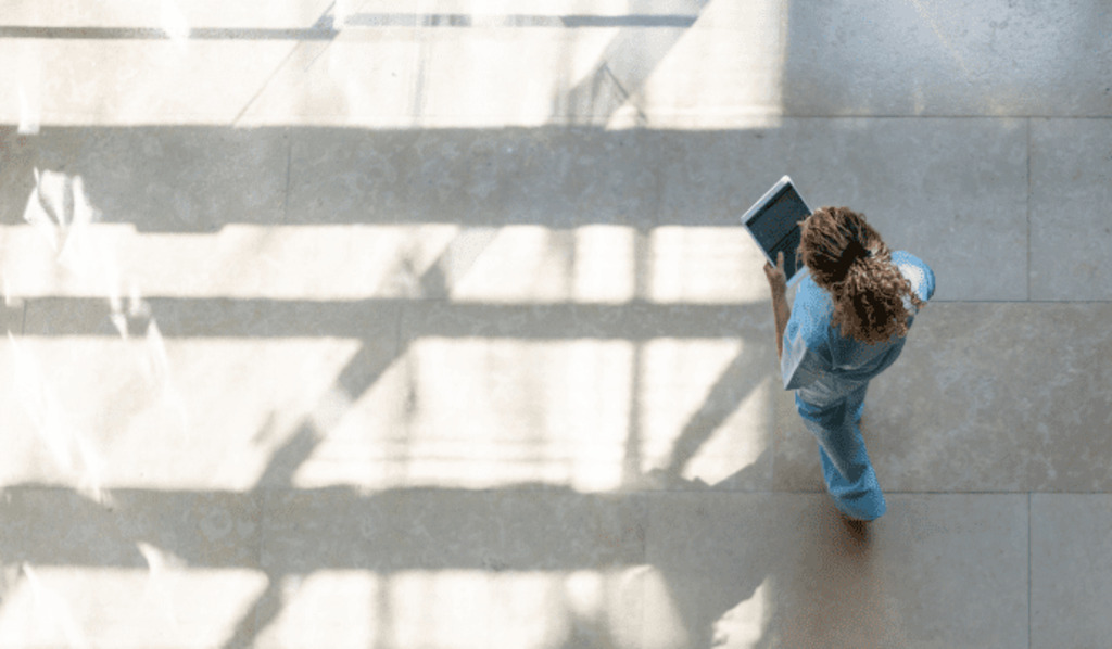 A person in light blue scrubs walks across a sunlit floor while holding a tablet, with shadows from windows casting patterns on the ground. The scene is viewed from above.