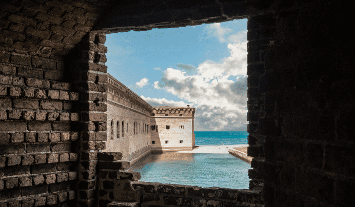 View of a historic brick fort and calm turquoise water, seen through a rectangular window in a brick wall, with a bright blue sky and fluffy clouds in the background.