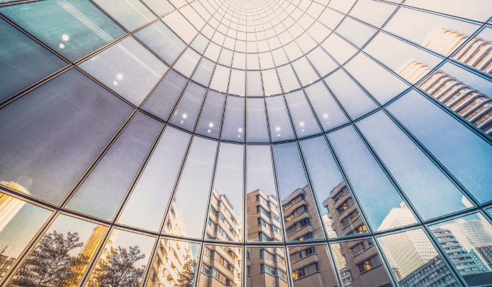 Looking up at a curved glass skyscraper facade reflecting surrounding buildings, trees, and a blue sky, creating a geometric, symmetrical pattern with a sense of depth and perspective.