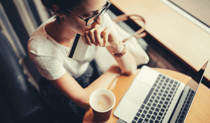 A woman wearing glasses sits at a table with a laptop, holding a credit card and looking thoughtful. A cup of coffee is in front of her, and sunlight streams in from a nearby window.