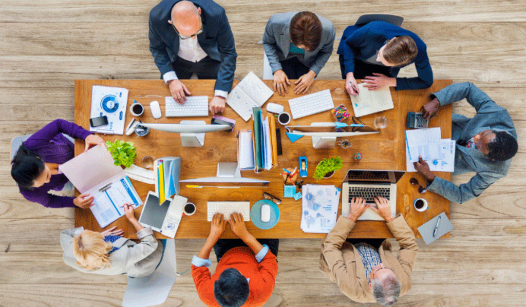 A group of eight people sit around a wooden table, working on laptops, tablets, and papers, engaging in discussion. The table is scattered with documents, coffee cups, and office supplies.