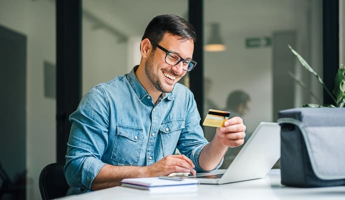 A smiling man wearing glasses and a denim shirt sits at a desk, holding a credit card in one hand and using a laptop with the other. A notebook and bag are on the table beside him.