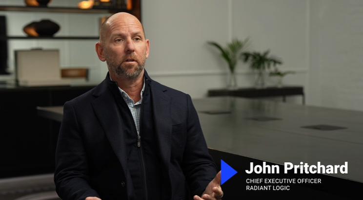 A man with a bald head and trimmed beard, wearing a dark suit jacket, sits and speaks in a modern office. Text on screen reads: John Pritchard, Chief Executive Officer, Radiant Logic.