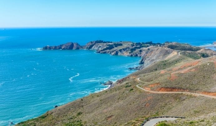 A winding road curves along a dry, grassy hillside overlooking a bright blue ocean, with rocky cliffs and a small peninsula jutting into the water beneath a clear sky.