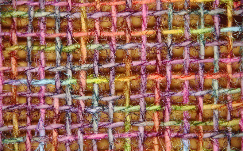 A close-up view of a colorful woven fabric, showing an intricate pattern of intersecting threads in pink, purple, orange, yellow, blue, and green, with visible texture and small gaps between the threads.